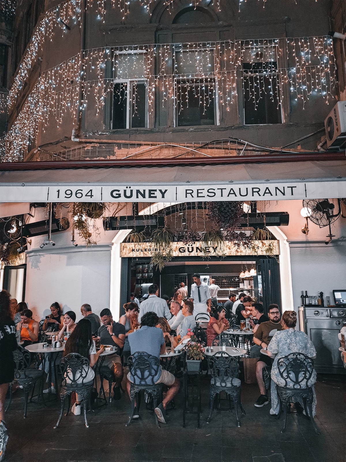 Evening crowd dining outside 1964 Güney Restaurant under strings of lights